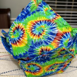 May include: A folded fabric bowl with a tie-dye pattern in shades of blue, green, yellow, and red. The bowl is made of cotton fabric and has a unique, handmade design.