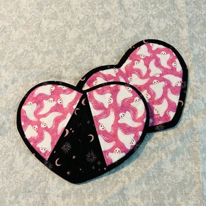 May include: Two heart-shaped pot holders with a pink and black fabric design. One side features a pink fabric with white ghost silhouettes. The other side features a black fabric with white stars and crescent moons.