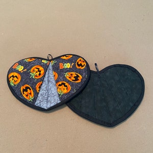 Puede incluir: Un par de agarraderas para ollas en forma de corazón. Una es negra con un reverso acolchado gris y la otra es negra con un patrón de calabazas naranjas y las palabras "Boo!" sobre un fondo gris.