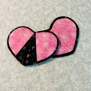 Peut inclure: Deux dessous de plat en forme de cœur. L'un est rose avec des toiles d'araignées blanches, l'autre est noir avec un croissant de lune et des étoiles. Les deux ont une bordure noire.