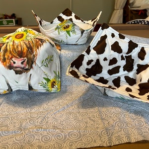 Puede incluir: Tres protectores de cuenco de tela. Uno presenta una vaca con girasoles, otro tiene un patrón de vaca y el tercero tiene un patrón de girasol.