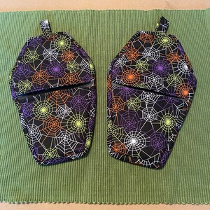 Puede incluir: Dos manoplas de horno negras con un patrón de telaraña en naranja, verde y morado. Las manoplas tienen forma de ataúdes.