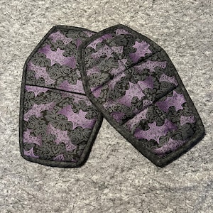 May include: Two black and purple fabric pot holders shaped like coffins with a bat pattern.