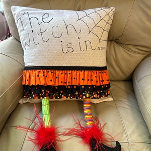 May include: Halloween-themed decorative pillow with the text "The witch is in..." and a spiderweb design. The pillow features orange and black ruffled trim, and witch legs with red feather accents and black shoes.