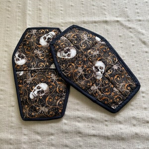 Peut inclure: Deux maniques en tissu noir et or en forme de cercueils avec un motif de crâne et de toile d'araignée.