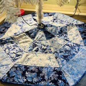 May include: A blue and white quilted Christmas tree skirt. The skirt is octagonal and features a patchwork design with floral and bird patterns. The color palette includes shades of blue and white.