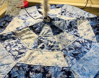 May include: A blue and white quilted Christmas tree skirt. The skirt is octagonal and features a patchwork design with floral and bird patterns. The color palette includes shades of blue and white.