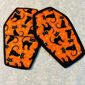 May include: Two orange and black oven mitts shaped like coffins with a black cat and gold star pattern.