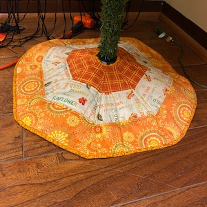 Op de afbeelding: Een ronde kerstboomrok in oranje en crèmekleurige tinten, met bloemen- en ruitpatronen. De rok heeft een geschulpte rand en tekst zoals "Sunflower" en "Harvest". Op een houten vloer.