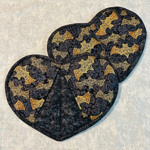 Puede incluir: Dos salvamanteles acolchados en forma de corazón. Los salvamanteles tienen un fondo negro con un estampado de murciélagos dorados. Los bordes están ribeteados con un ribete negro. Para uso en la cocina.