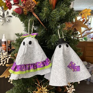 Può includere: Due ornamenti a forma di fantasma fatti a mano con tessuto grigio a spirali. Uno ha una balza viola e verde con stelle gialle, l'altro un cappello nero e toppe viola con stelle gialle. Entrambi hanno occhi neri a bottone e sono appesi con spago.