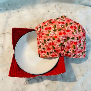 May include: A white ceramic plate with a light blue rim sits on a red quilted square. A cherry-patterned bowl cozy covers the plate. The cozy features red cherries, green leaves, and white flowers on a pink gingham background. The plate and cozy are on a marble surface.