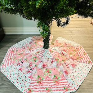 May include: A festive Christmas tree skirt with a quilted design. The skirt has a pink and white colour scheme, featuring snowmen, poinsettias, and candy canes. The octagonal skirt is made of fabric.