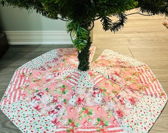Könnte beinhalten: Ein gesteppter Weihnachtsbaumrock mit festlichem Design. Der Rock hat ein rosa-weißes Farbschema mit Mustern von Schneemännern, Weihnachtssternen und Zuckerstangen. Der Rock ist achteckig und aus Stoff.