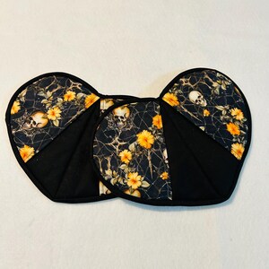 Peut inclure: Deux maniques noires avec un imprimé floral. L'imprimé floral présente des fleurs jaunes, des crânes et des toiles d'araignées. Les maniques ont une forme unique et un bord noir.