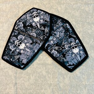May include: Two black and white fabric pot holders shaped like coffins with a skull and floral design.