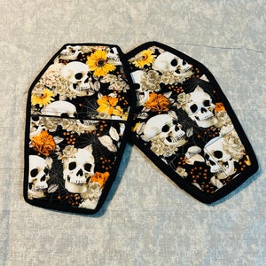 May include: Two coffin-shaped oven mitts with a black border. The fabric features a pattern of white skulls, orange and yellow flowers, and spiderwebs on a black background. A Halloween-themed kitchen accessory.