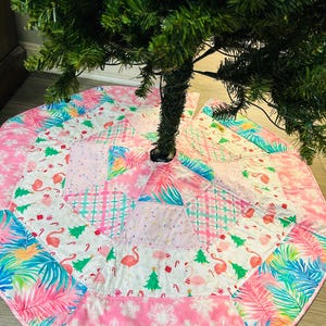 May include: A pink and white Christmas tree skirt with a tropical flamingo and palm leaf pattern. The skirt has a patchwork design with a snowflake border and is placed under a green Christmas tree.