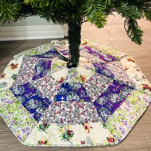May include: A quilted Christmas tree skirt with a purple, white, and green floral pattern. The skirt features the words "He is Risen" and "Hope" in a white font.
