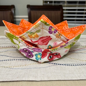 May include: A quilted orange and white fabric bowl with a floral and cat print. The bowl is folded into a star shape.