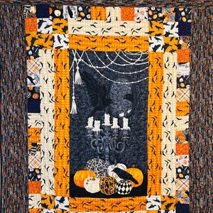 May include: A Halloween-themed quilt with a central panel featuring a black raven, pumpkins, and a candelabra. The quilt has a patchwork design with orange, black, and white fabrics, including bat and skull patterns. The quilt is held by red clothespins.
