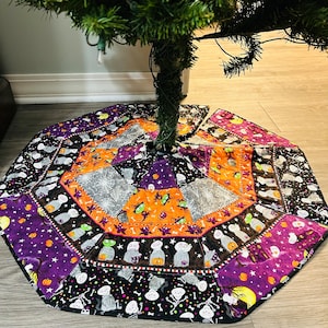 May include: A Halloween-themed tree skirt with a patchwork design. The skirt features various Halloween motifs, including ghosts, skeletons, spiders, and pumpkins, in orange, purple, black, and gray colors. The skirt is placed around the base of a green Christmas tree.
