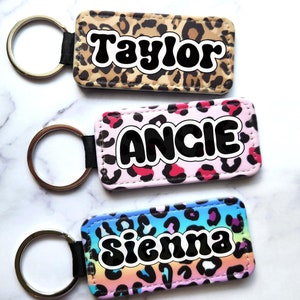 May include: Three keychains with leopard print fabric and different colored backgrounds. The keychains have the names "Taylor", "Angie", and "Sienna" written in black, bubble letters.