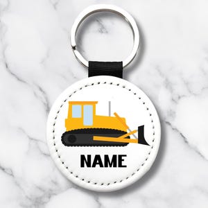 May include: A round white keychain with a black strap and silver ring. The keychain features a yellow and black bulldozer graphic with the word "NAME" in black block letters. The background is a marble pattern.