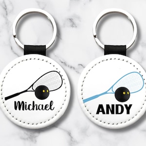 May include: Two white round keyrings with a black strap and silver ring. Each keyring features a squash racket and ball design. One keyring has the name "Michael" and the other has the name "ANDY" in black text.