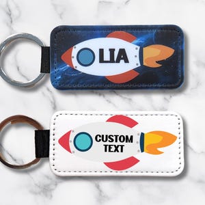 May include: Two rectangular keychains with rocket ship designs. One has a blue background with the name "LIA", the other has a white background with "CUSTOM TEXT". Both have a silver or bronze key ring.