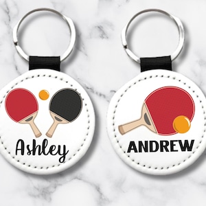 May include: Two white round keychains with black key rings. Each keychain has a different name, Ashley and Andrew, and a ping pong paddle and ball design.