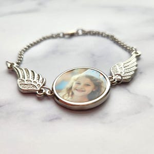 Może przedstawiać: Srebrna bransoletka z okrągłym wisiorkiem na zdjęcie przedstawiającym małą dziewczynkę w koronie. Bransoletka ma po obu stronach wisiorka dwa srebrne skrzydła anioła.
