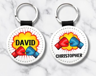 Puede incluir: Dos llaveros de cuero blanco con anillas de llaves negras. Cada llavero tiene una etiqueta redonda de cuero blanco con un gráfico de explosión de estilo cómic amarillo y rojo. Una etiqueta dice "DAVID" y la otra dice "CHRISTOPHER".