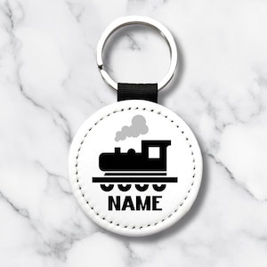 May include: A white, round keychain with a black train graphic and the word "NAME" printed below. The keychain has a silver ring and a black strap. The train graphic includes a smokestack with gray smoke.