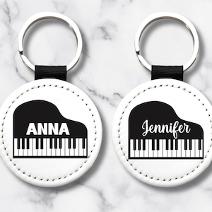 May include: Two white keychains with a piano design. Each keychain features a black piano graphic with a name, "ANNA" and "Jennifer", in a bold font. The keychains have a silver ring and a black strap.