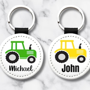 Könnte beinhalten: Zwei runde Schlüsselanhänger mit weißem Hintergrund und schwarzem Riemen. Einer hat ein grünes Traktormotiv mit dem Namen "Michael" in Schwarz. Der andere hat ein gelbes Traktormotiv mit dem Namen "John" in Schwarz.