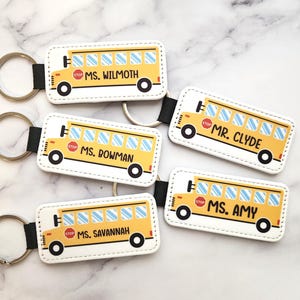 May include: Four yellow school bus keychains with different teacher names. Each keychain has a white background with black stitching around the edges. The keychains are attached to silver key rings.