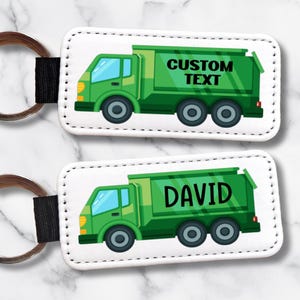Op de afbeelding: Twee witte rechthoekige sleutelhangers met een groen cartoon vuilniswagen ontwerp. Eén sleutelhanger heeft de tekst "CUSTOM TEXT" en de andere de naam "DAVID" in het groen. Elke sleutelhanger heeft een zwarte band en een zilveren sleutelring.