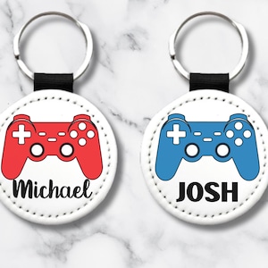 Peut inclure: Deux porte-clés ronds blancs avec des anneaux de clés noirs. Chaque porte-clés présente un fond blanc avec un contour noir d'une manette de jeu vidéo. Une manette est rouge et l'autre est bleue. Le porte-clés avec la manette rouge porte le nom "Michael" imprimé en dessous. Le porte-clés avec la manette bleue porte le nom "Josh" imprimé en dessous.
