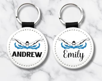 Puede incluir: Dos llaveros redondos blancos con anillos de llaves negros. Cada llavero presenta una silueta en blanco y negro de un nadador haciendo el estilo mariposa con agua azul. El texto "Andrew" está en un llavero y "Emily" en el otro.
