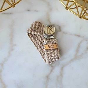 以下が含まれることがあります： ベージュのかぎ針編みのおしゃぶりクリップ。金属製の留め金と「Same design」と書かれた小さな木製のタグが付いています。 クリップは大理石の表面にあり、背景には2つの金色の幾何学的なキャンドルホルダーがあります。