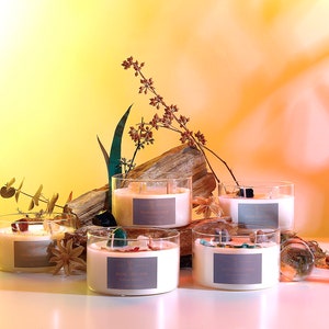May include: Five glass candle jars with natural wax and crystals inside. The candles are arranged on a wooden surface with greenery and crystals around them. The background is a gradient of yellow and orange.