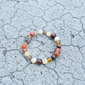 May include: A bracelet made of various colored stones, including red, orange, brown, and white. The bracelet has a gold clasp and is lying on a cracked gray surface.