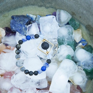 Może przedstawiać: Bransoletka z czarnych koralików z kwadratowym złotym urokiem zawierającym czarną kulę lawy. Bransoletka jest otoczona lodem i różnokolorowymi kamieniami szlachetnymi.