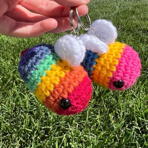 May include: Two crocheted rainbow bee keychains with white wings and black eyes. The keychains are attached to a silver key ring.