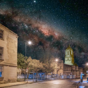 May include: A night view of a street in a city with a church tower in the distance. The Milky Way is visible in the night sky above the buildings.