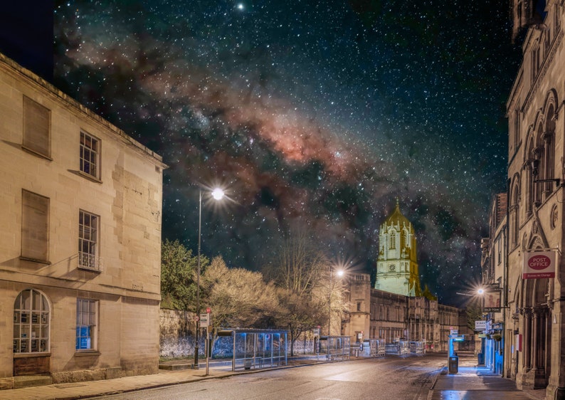 May include: A night view of a street in a city with a church tower in the distance. The Milky Way is visible in the night sky above the buildings.