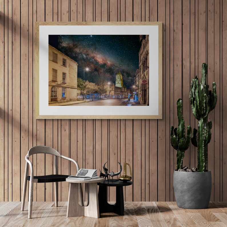 May include: A framed photograph of a city street at night with a view of the Milky Way in the sky. The street is lined with buildings and streetlights. The photo is hanging on a wooden wall.