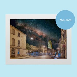 May include: A night time photograph of a street in a city with a church steeple in the distance. The Milky Way is visible in the night sky above the street.