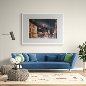 May include: A blue sofa with green throw pillows sits in a living room with a framed photograph of a city street at night with the Milky Way visible in the sky above. The sofa is in front of a white rug with a geometric pattern. A floor lamp with a black shade stands to the left of the sofa. A potted plant is on the right side of the sofa.